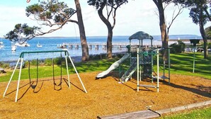 Ein Spielplatz mit Schaukeln und Rutsche, nahe eines Gewässers mit Booten und einem Steg im Hintergrund.