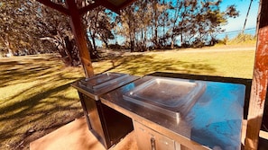 An outdoor kitchen with a sink and a grill, surrounded by trees and a clear sky.