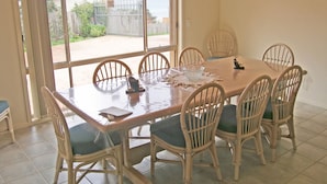 A dining area with a glass-top table, six wicker chairs with cushions, and a view of a garden through large windows.