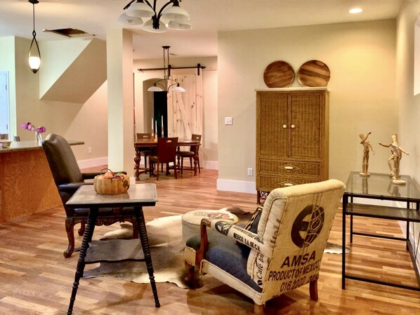 A spacious living room with a wooden floor, a dining area with a table and chairs, a large cabinet with decorative plates, and a unique armchair with a label. - The apARTment/ historic railroad inn, pet-friendly (Cowan)