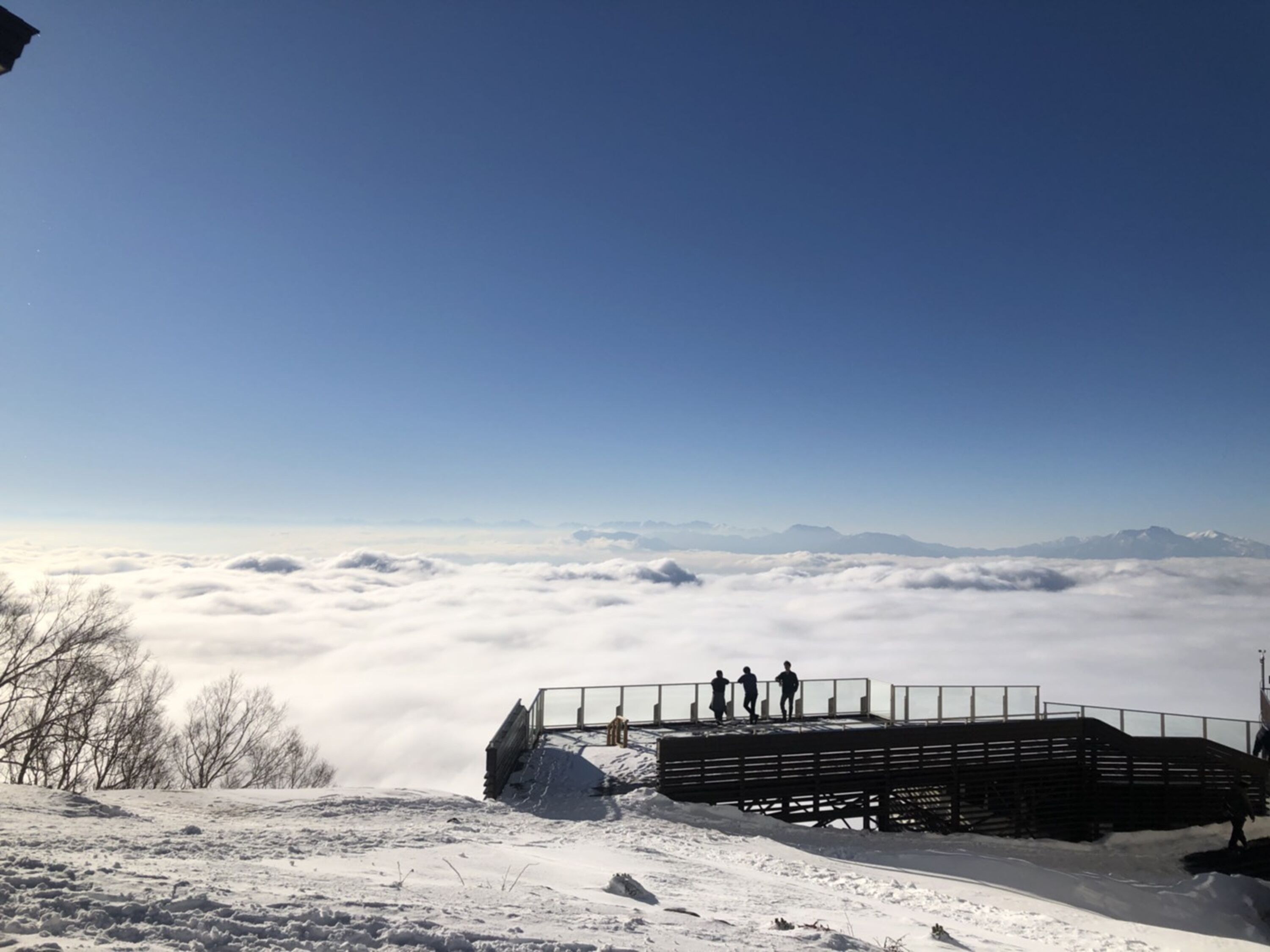 A snow-covered landscape with a wooden viewing platform and a clear view of the clouds and distant mountains.