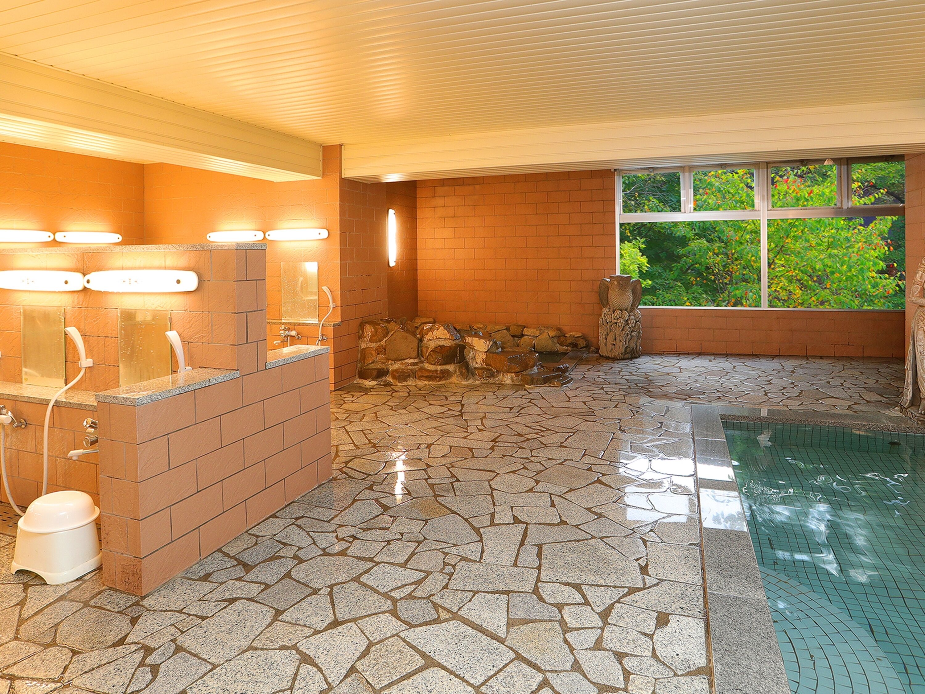 A bathroom with a stone floor, a sink with lights, a window with a view of greenery, and a stone wall.