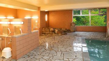 A bathroom with a stone floor, a sink with lights, a window with a view of greenery, and a stone wall.
