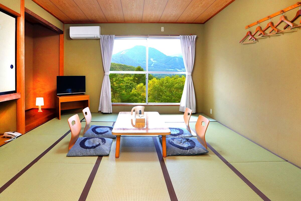 Habitació japonesa tradicional amb sòl de tatami, una taula baixa amb coixins, una televisió i vista a les muntanyes a través d'una finestra gran.