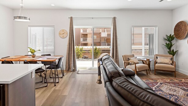 A modern living room with a leather sofa, dining table, and chairs. There is a large window with blinds, a ceiling fan, and a view of a building outside. - St. George Getaway, Brand New 5 BD Luxury Townhome (St. George)