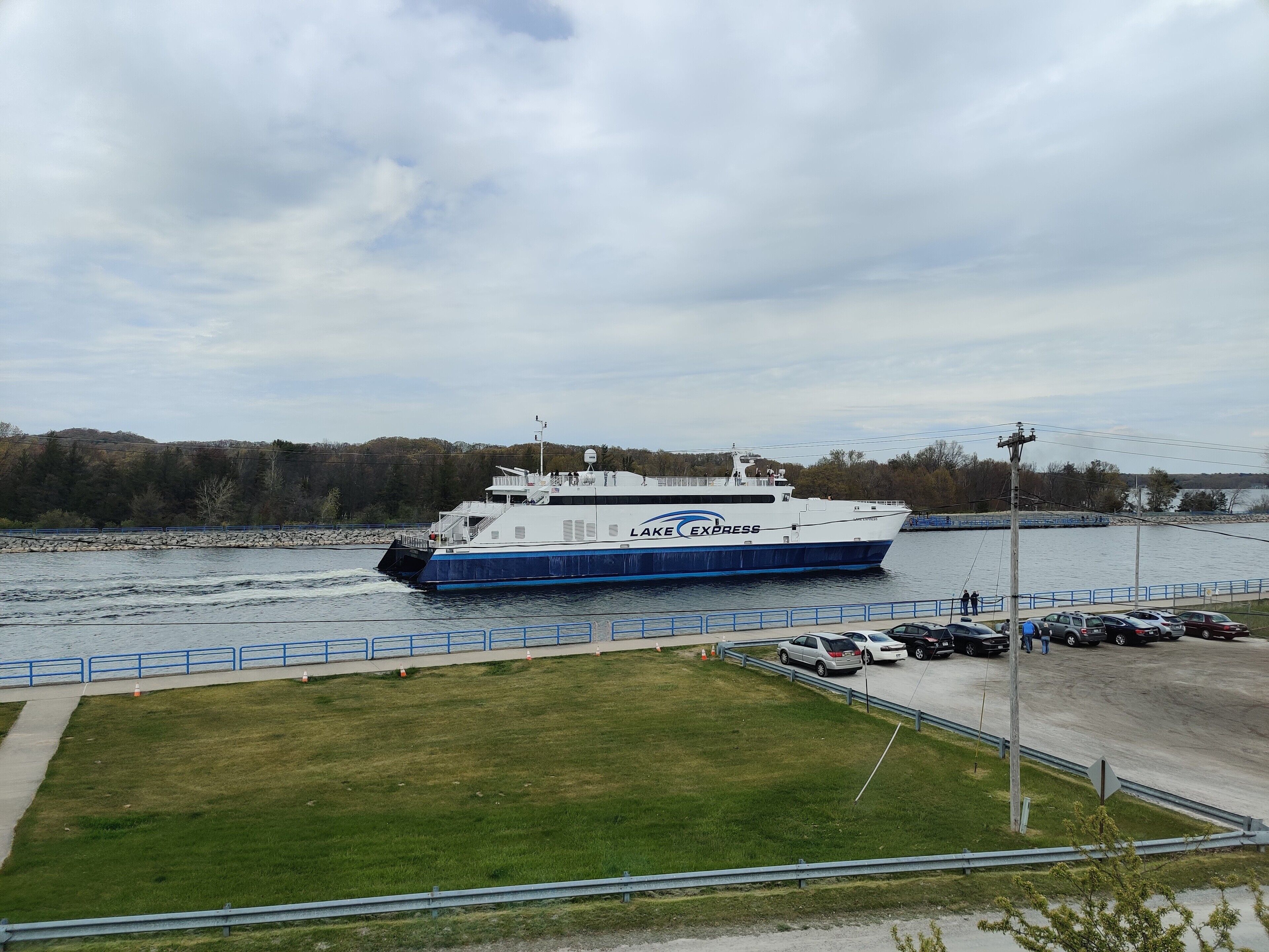 Un ferry atracado en un muelle con un área verde y coches aparcados cerca.