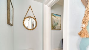 Un pasillo con un espejo redondo, un cuadro enmarco, una mesita blanca y un tapiz de macramé en la pared.