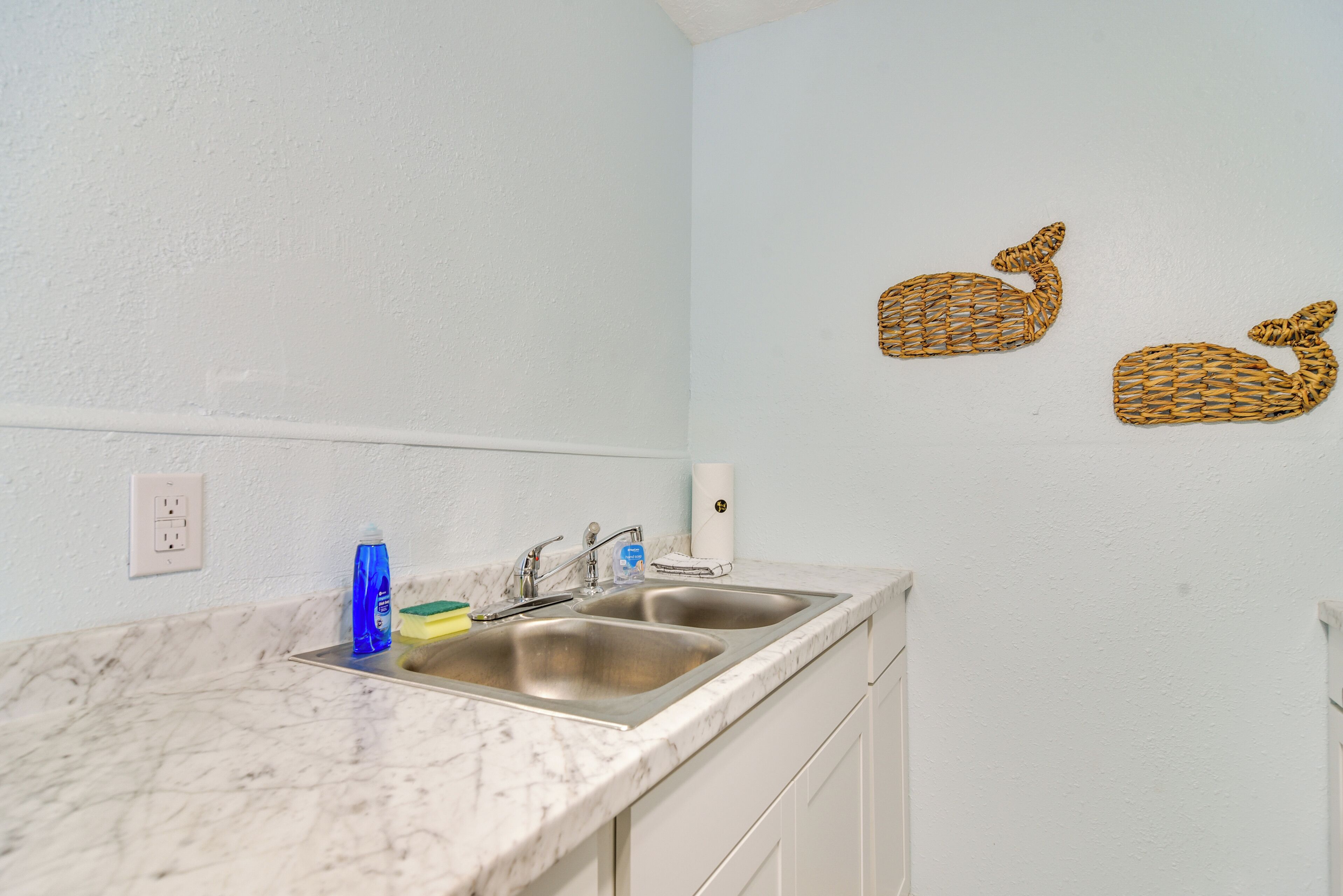 A kitchen with two stainless steel sinks, a marble countertop, and a white wall with two woven wall art pieces.