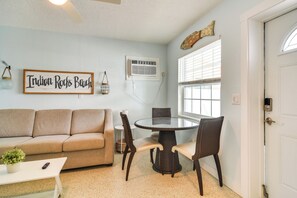 Uma sala de estar com um sofá bege, uma mesa de jantar redonda com cadeiras, uma pequena mesa branca com uma planta e um ar-condicionado fixado na parede.