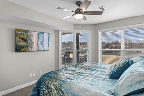 A bedroom with a bed, a ceiling fan, a painting, and a view through the window.