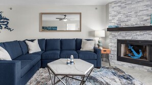 Un salon comprenant un canapé d’angle, une table basse en marbre, une cheminée décorée d’une ancre bleue et un ventilateur de plafond.