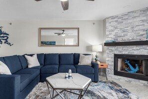 A living room with a sectional sofa, a marble coffee table, a fireplace with a blue anchor decoration, and a ceiling fan.