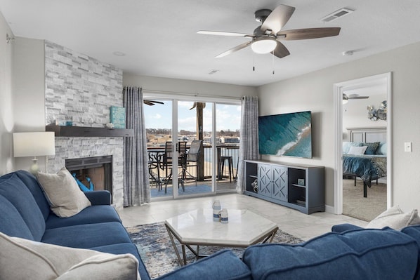 A modern living room with a fireplace, a ceiling fan, and a large window overlooking a balcony.