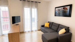 Un salon avec un canapé gris, un bureau en bois, une télévision et un tableau représentant des bateaux.