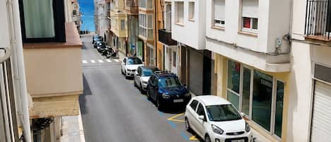 A narrow street lined with multi-story residential buildings, parked cars, and a clear view of the sea.