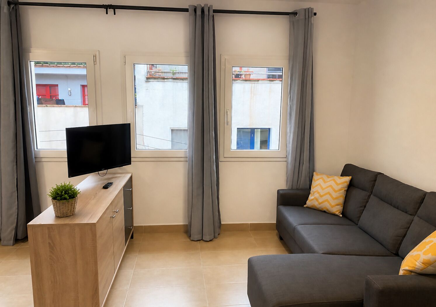 Ein modernes Wohnzimmer mit einer Couch, einem Fernseher auf einem Holztisch und zwei mit Vorhängen versehenen Fenstern.