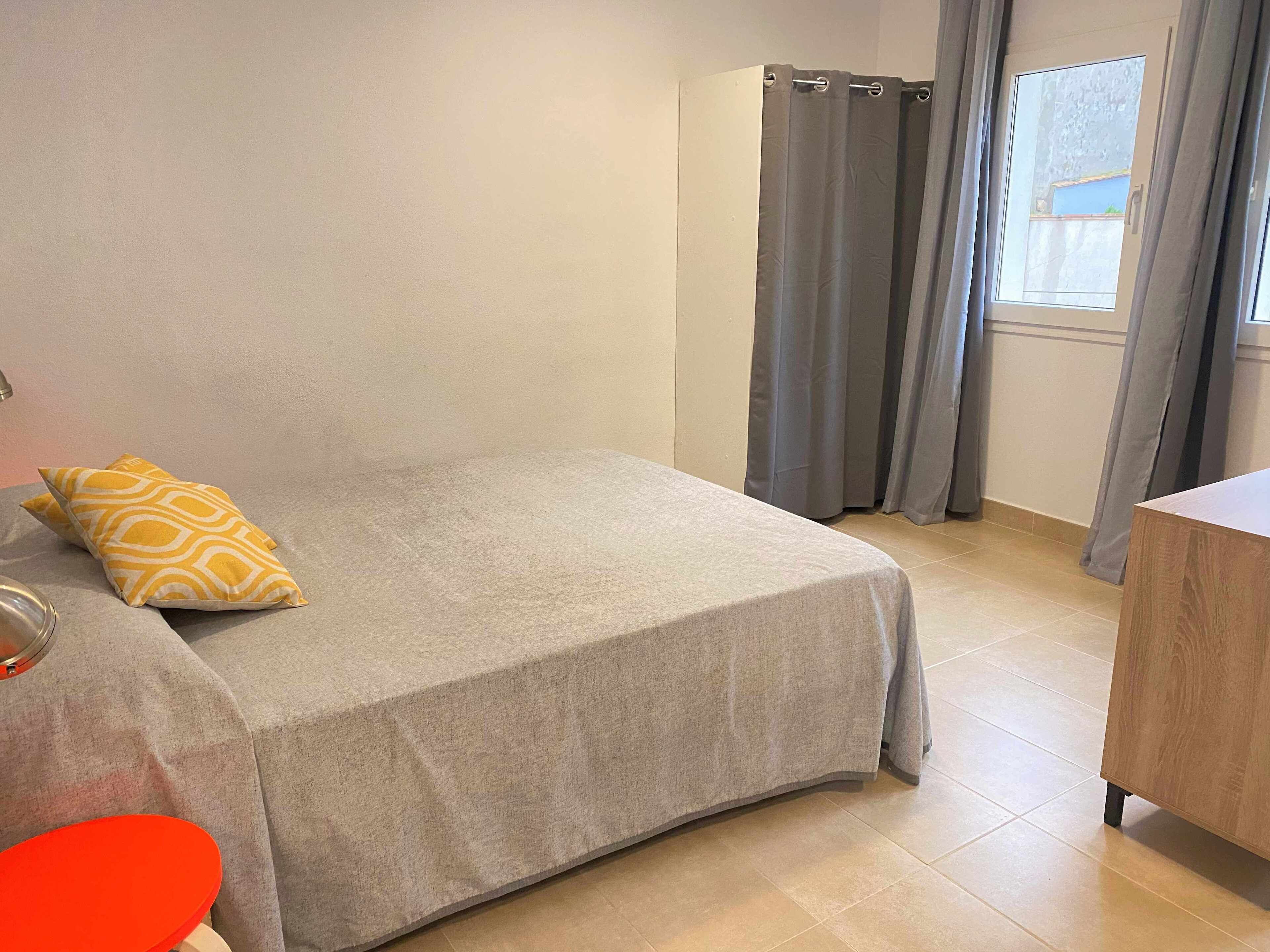 Un lit bien fait, recouvert d’une couverture grise et agrémenté d’un seul oreiller jaune, une table de chevet en bois avec une chaise rouge, et une fenêtre ornée de rideaux gris.