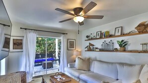 Una sala de estar con ventilador de techo, un sofá, una mesa de centro y un televisor. Hay una puerta corrediza de vidrio que da a un espacio al aire libre.