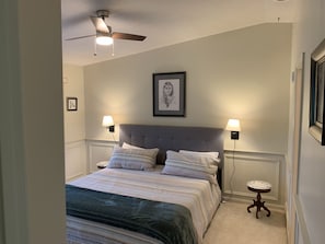 Een slaapkamer met een bed, twee bedlampen, een plafondventilator, een ingelijst kunstwerk aan de muur en een klein rond tafeltje naast het bed.