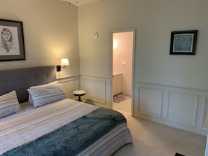 Un dormitorio con una cama, una mesita de noche, una lámpara, un cuadro enmarcado y una puerta que da al baño.