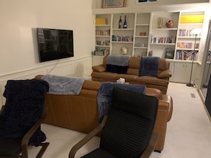 Een woonkamer met een bank, een fauteuil en een televisie. Er staan boekenkasten met boeken en decoratieve objecten.