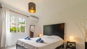 Ein Schlafzimmer mit Bett, Nachttischlampe, kleinem Tisch mit Lampe, Fenster mit Vorhängen und einer Pendelleuchte.
