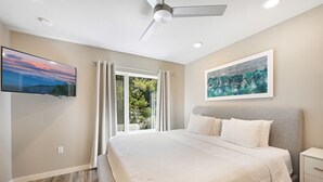 Una camera da letto con un letto, un ventilatore a soffitto, una TV, una finestra con tende e un quadro appeso al muro.