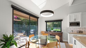 Eine moderne Küche mit einem Glastisch, weißen Schränken und einem großen Fenster mit Blick auf einen Garten.