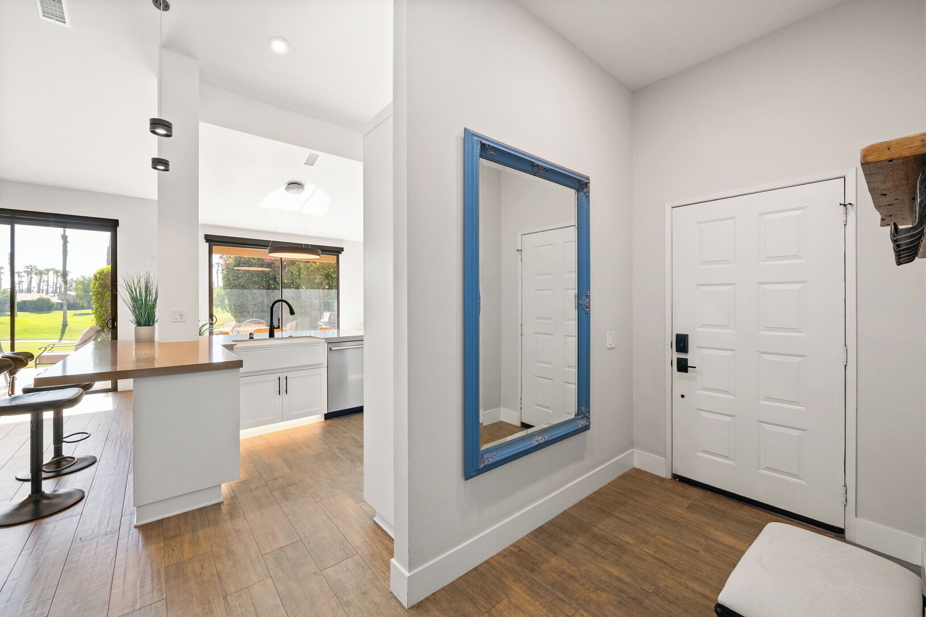 A modern kitchen with white cabinets, a large island with bar stools, a blue-framed mirror, and a wooden floor.