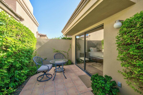 A patio with two chairs and a table, surrounded by greenery and a glass door leading to an interior space.