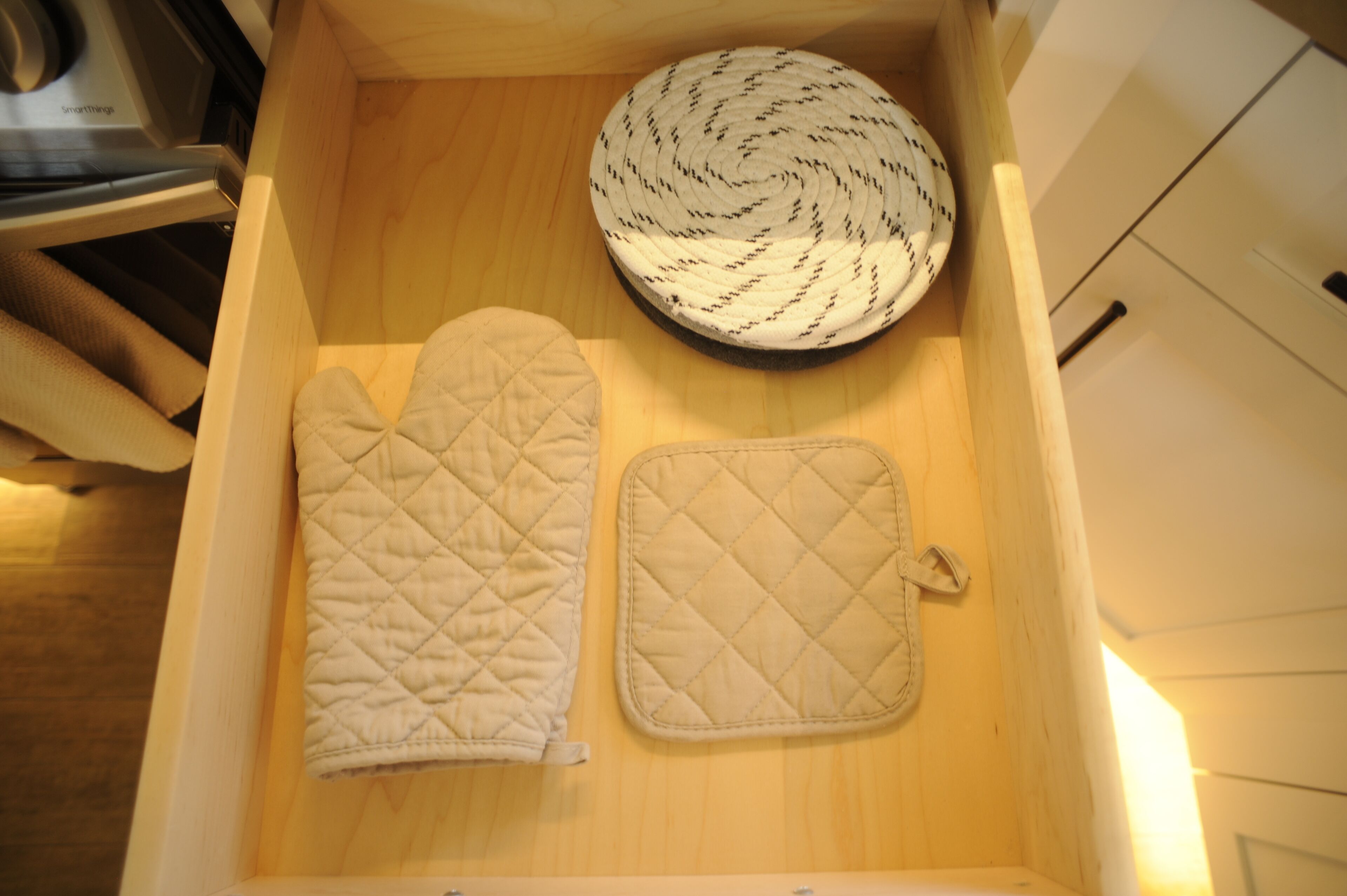 A kitchen drawer with a wooden interior, containing a white oven mitt, a white pot holder, and a woven kitchen towel.