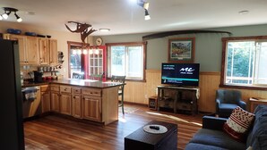 A modern kitchen with wooden cabinets, a central island, and a dining area with a table and chairs. A flat-screen TV is mounted on the wall, and there is a window providing natural light. - Resurrection Peaks Lodge.  Upper unit sleeps 6, private lot and close to town. (Seward)