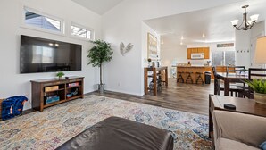 A spacious living area with a large flat-screen TV, a coffee table, and a sofa. There is a kitchen in the background with wooden cabinets and a bar area. - Cozy House Near Boise State University, Downtown and Boise River Greenbelt. (Boise)
