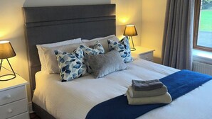 Una cama bien hecha con un edredón azul, un almohadón gris y un cojín estampado. Hay dos lámparas de noche sobre mesitas blancas.