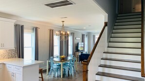 A modern kitchen with white cabinets, a dining area with blue chairs and a chandelier, and a wooden staircase.