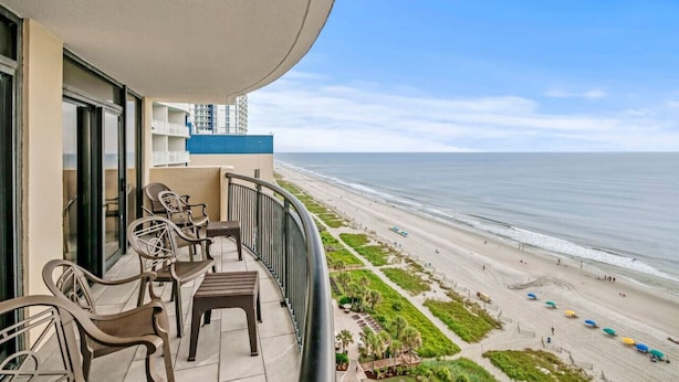 A balcony with chairs and a table overlooking a beach with umbrellas and people. - Oceanfront 3BR/2BA,walking distance to everything. (Myrtle Beach)