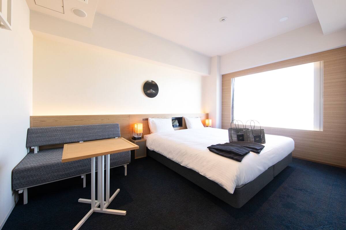 A modern hotel room with a bed, a desk, a chair, and a large window.
