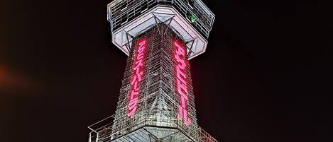 Uma torre alta e iluminada com ponta, rodeada por um edifício com vários andares e telhado plano.