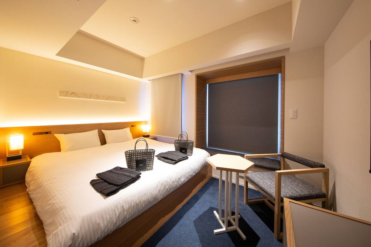 A hotel room with a large bed, bedside lamps, a small table with a chair, and a window with a view.