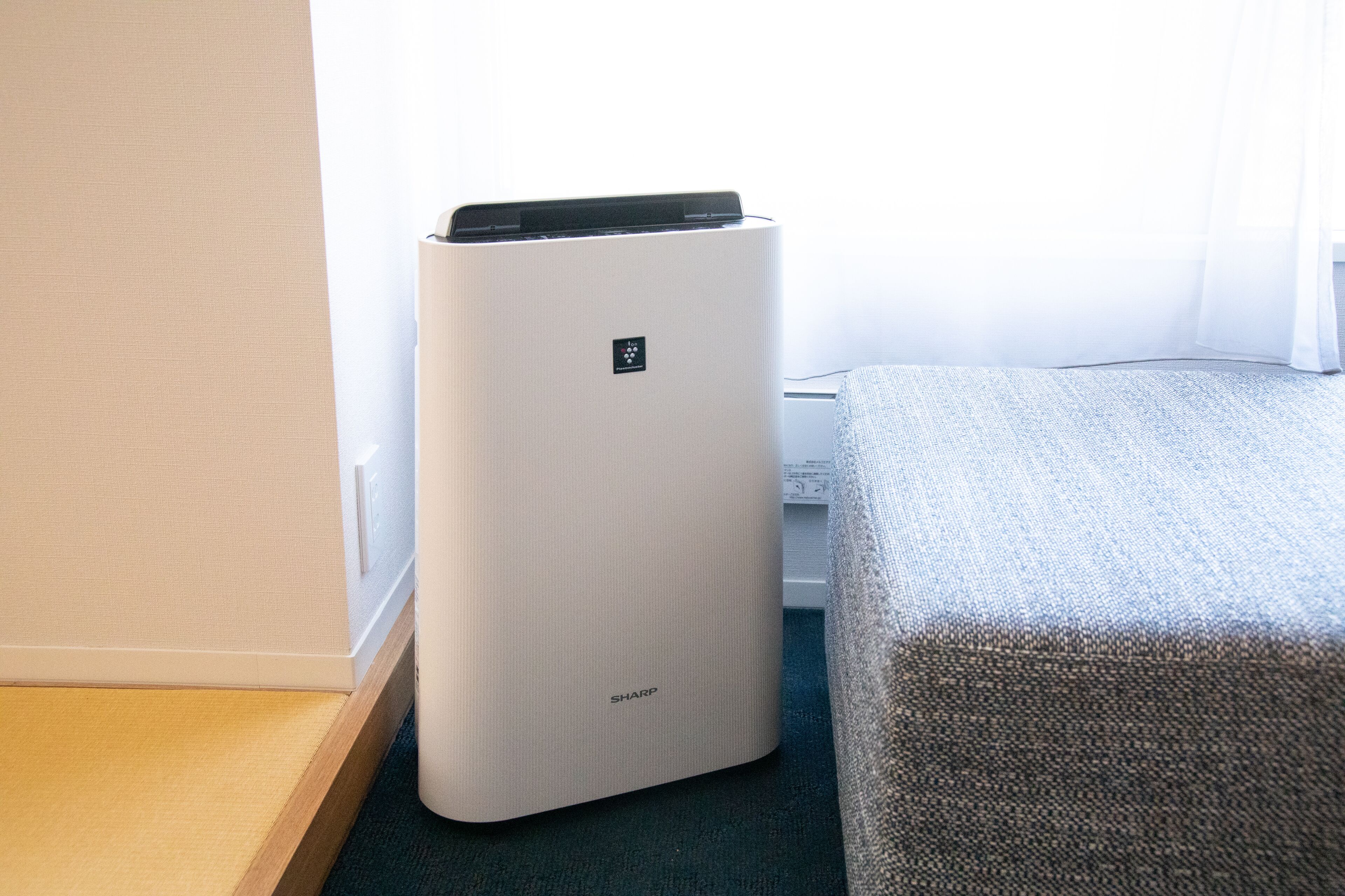 A room with a white air purifier, a bed with a grey cover, and a window with sheer curtains.