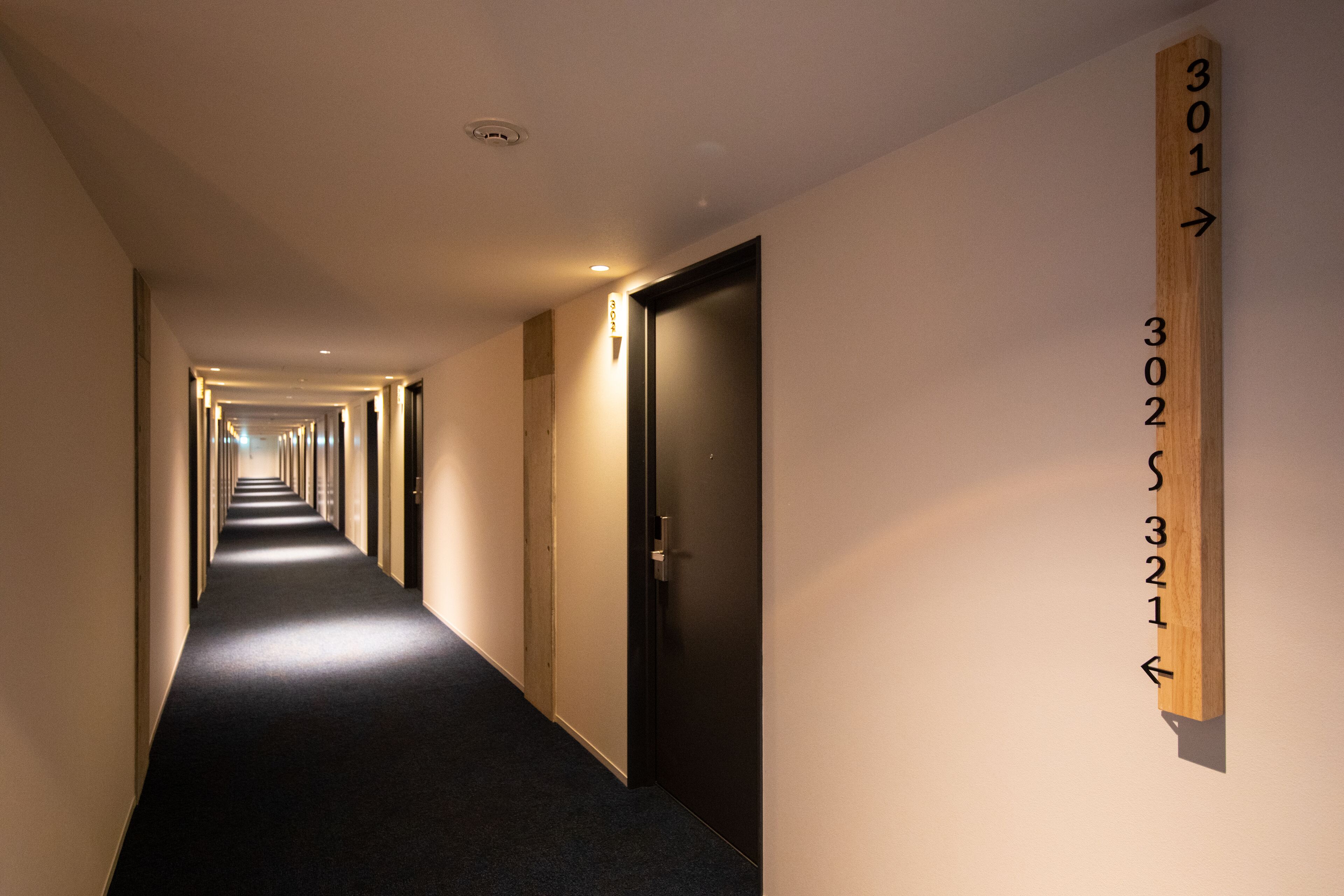 A hotel hallway with a carpeted floor, a wooden sign with room numbers, and doors on the right.