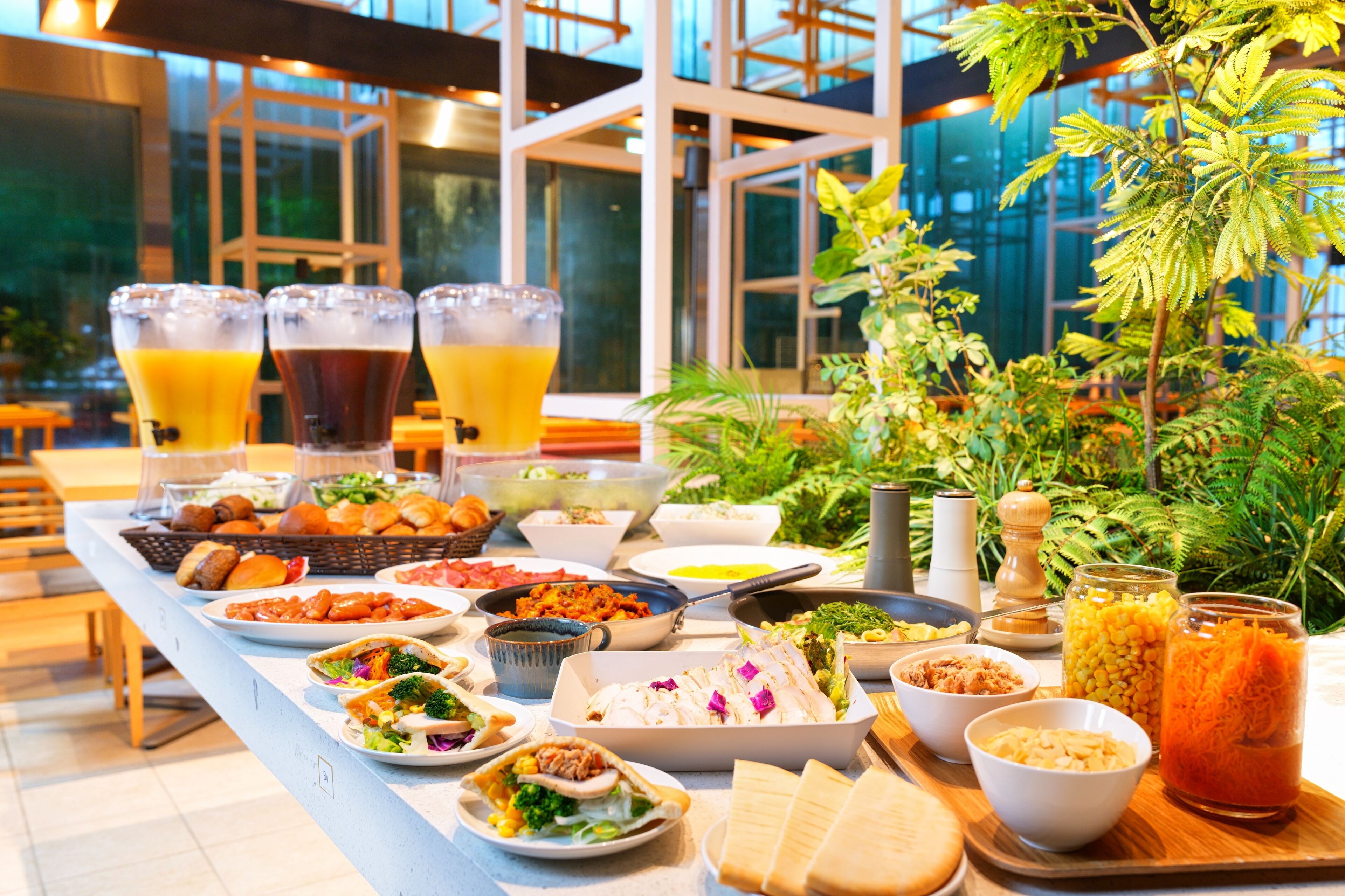 Una mesa de buffet con diversos platos y bebidas, rodeada de plantas de interior y amplios ventanales.