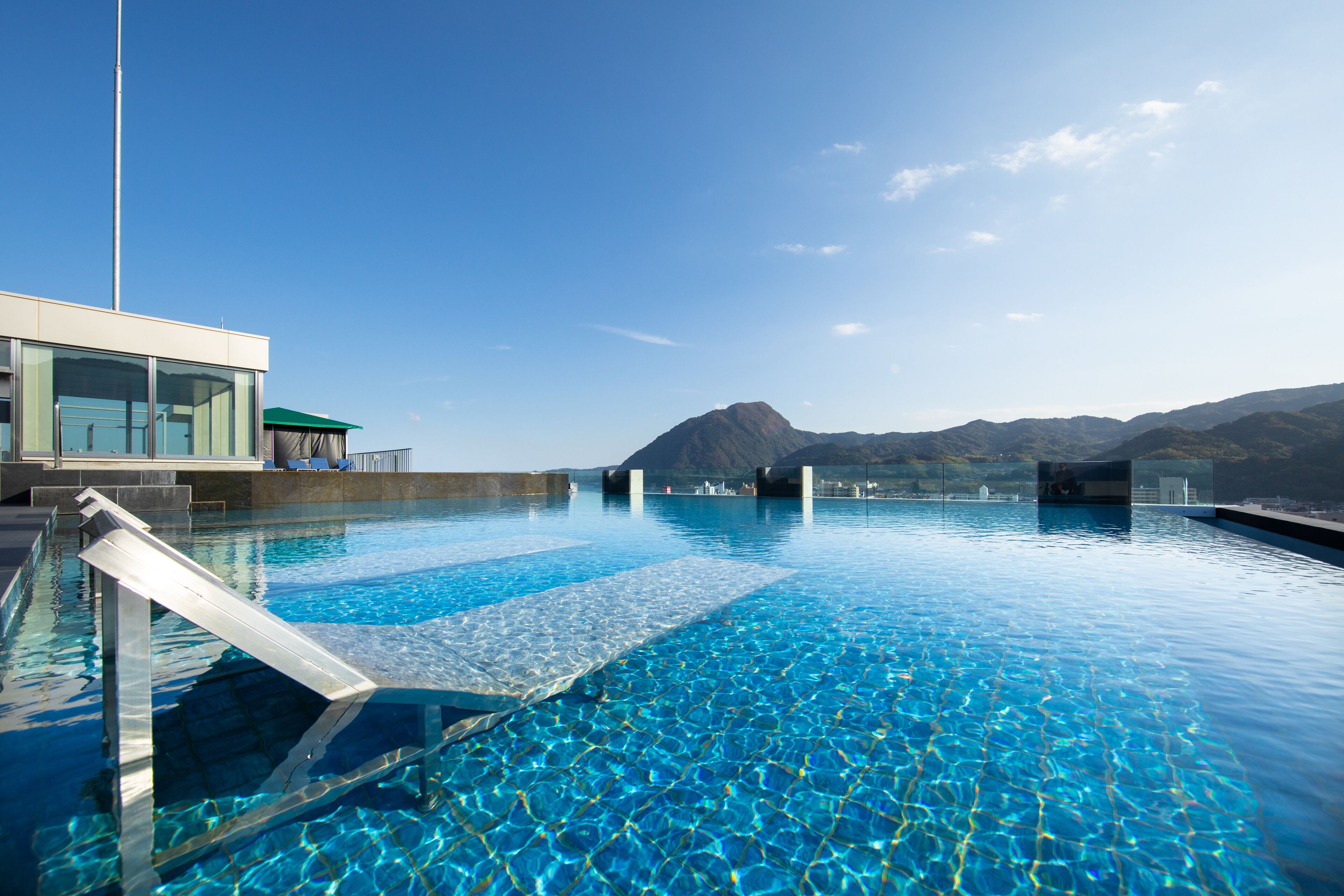 A clear blue pool with a glass edge, surrounded by a modern building with large windows and a mountain view in the background.