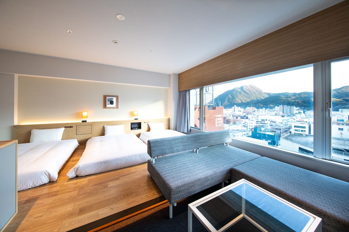 A hotel room with two beds, a sofa, a small table, and a city view through large windows.