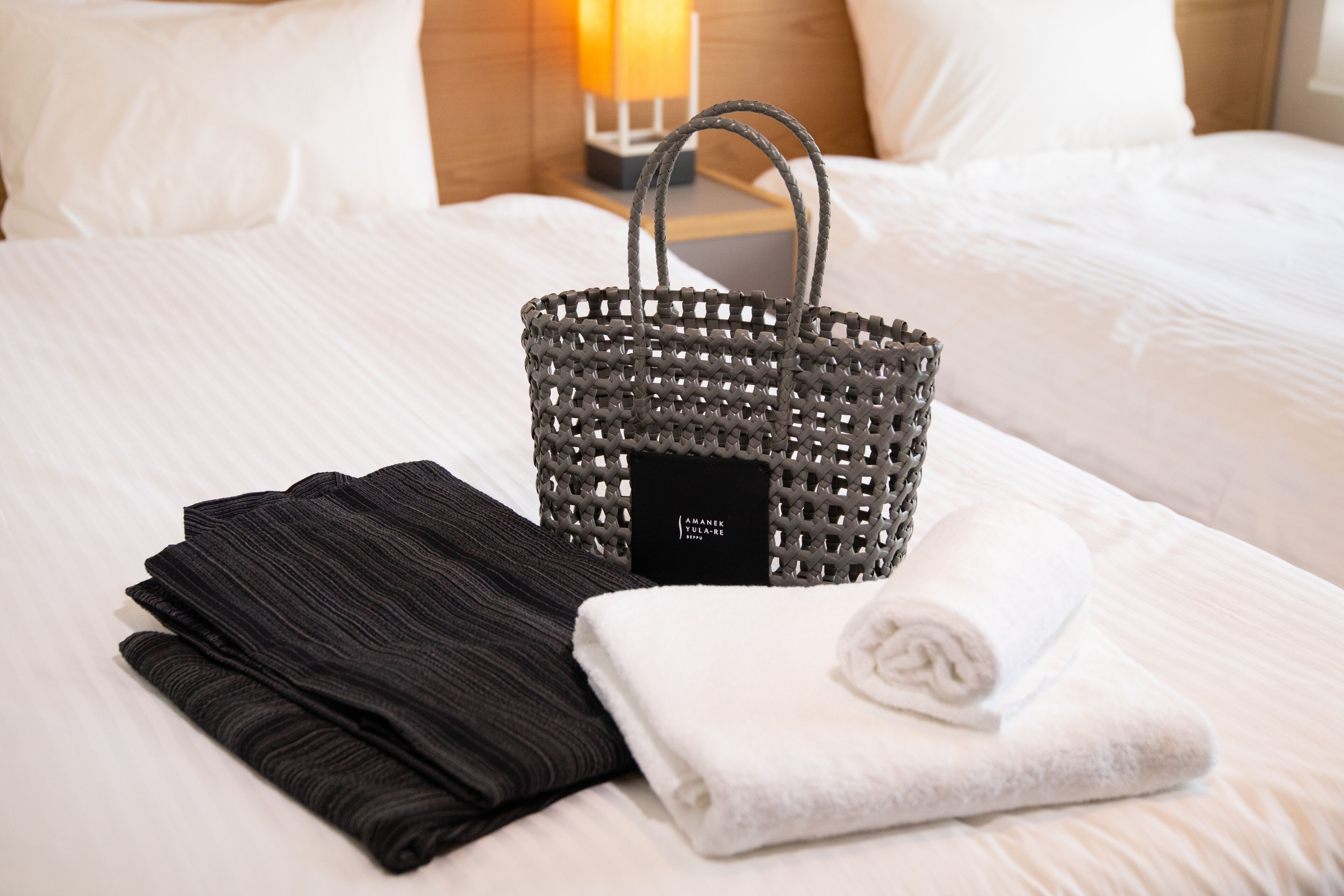 Una cama bien hecha con una toalla negra doblada, una toalla blanca enrollada y una cesta tejida con una etiqueta en una habitación de hotel.