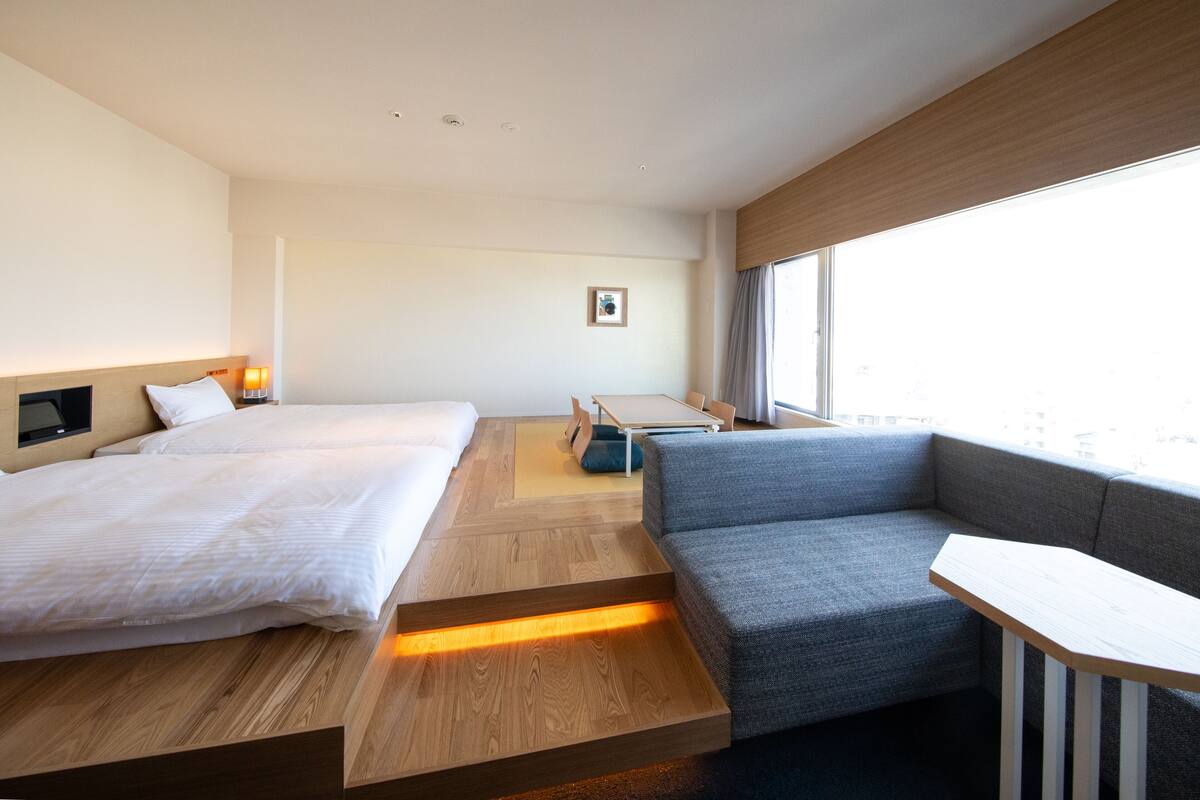 A modern hotel room with a wooden floor, a large window, a sofa, a bed, and a small table.