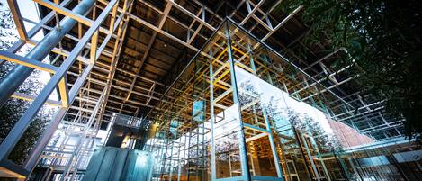 A modern building with a glass facade and metal framework, featuring an open interior space with tables and chairs.