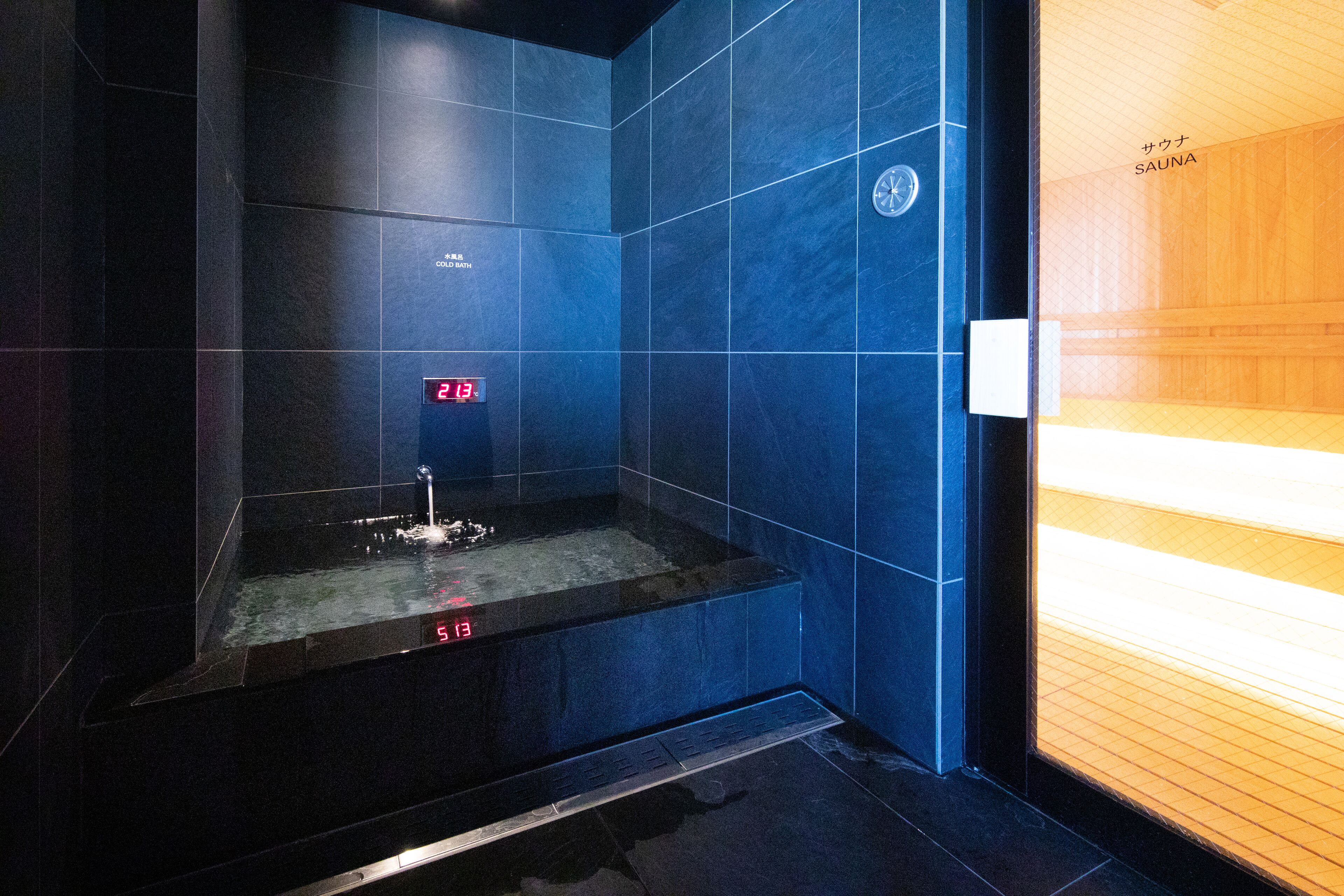 A modern sauna with dark blue tiles, a digital clock, and a wooden panel on the right.