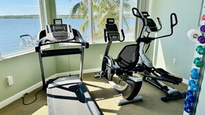 Et fitnessrum med løbebånd og crosstrainere, store vinduer med udsigt over havet og palmer.