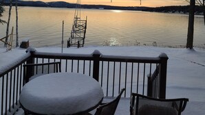 Ein mit Schnee bedeckter Deck mit Tisch und Stühlen, mit Blick auf einen stillen See bei Sonnenuntergang.
